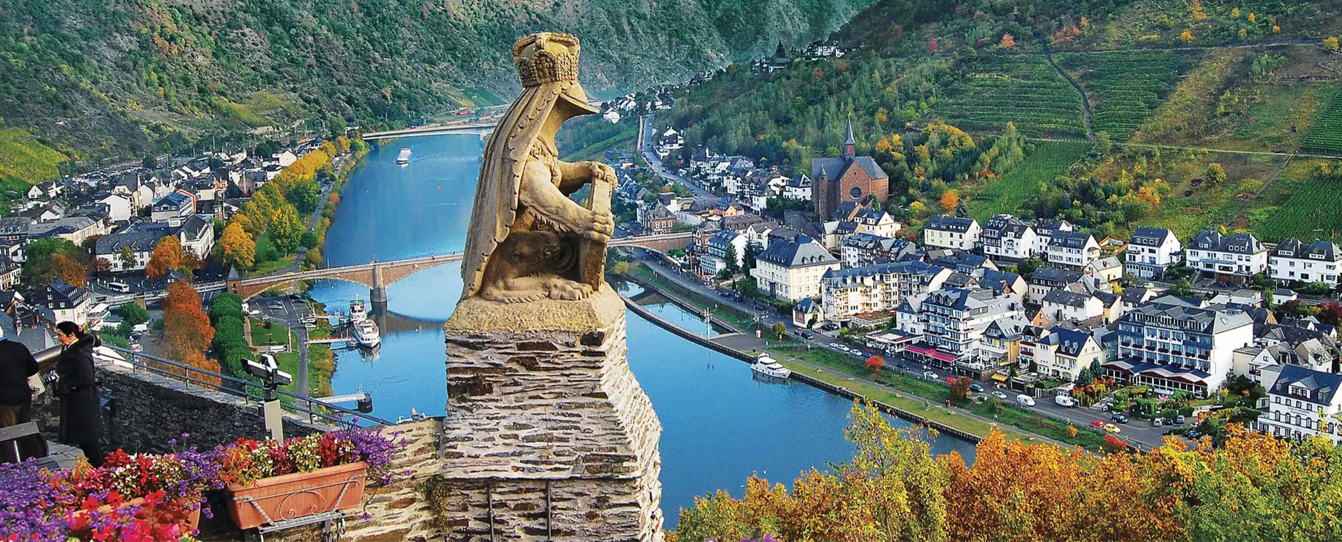 A statue overlooks a river and a town nestled between green hills. The sky is blue.