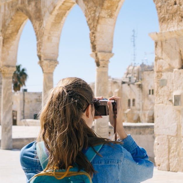 Woman taking a photo under stone arches in a sunlit historic setting.