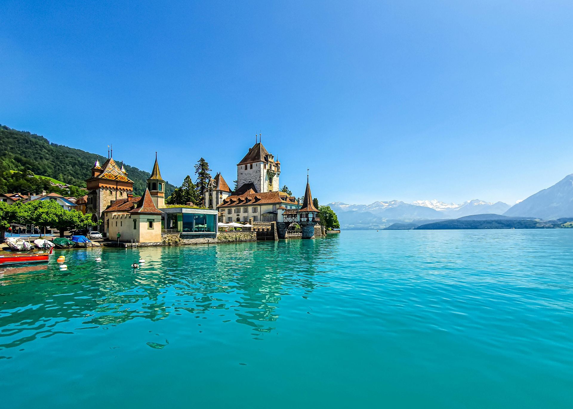 Castle on turquoise lake with mountains in background under blue sky.