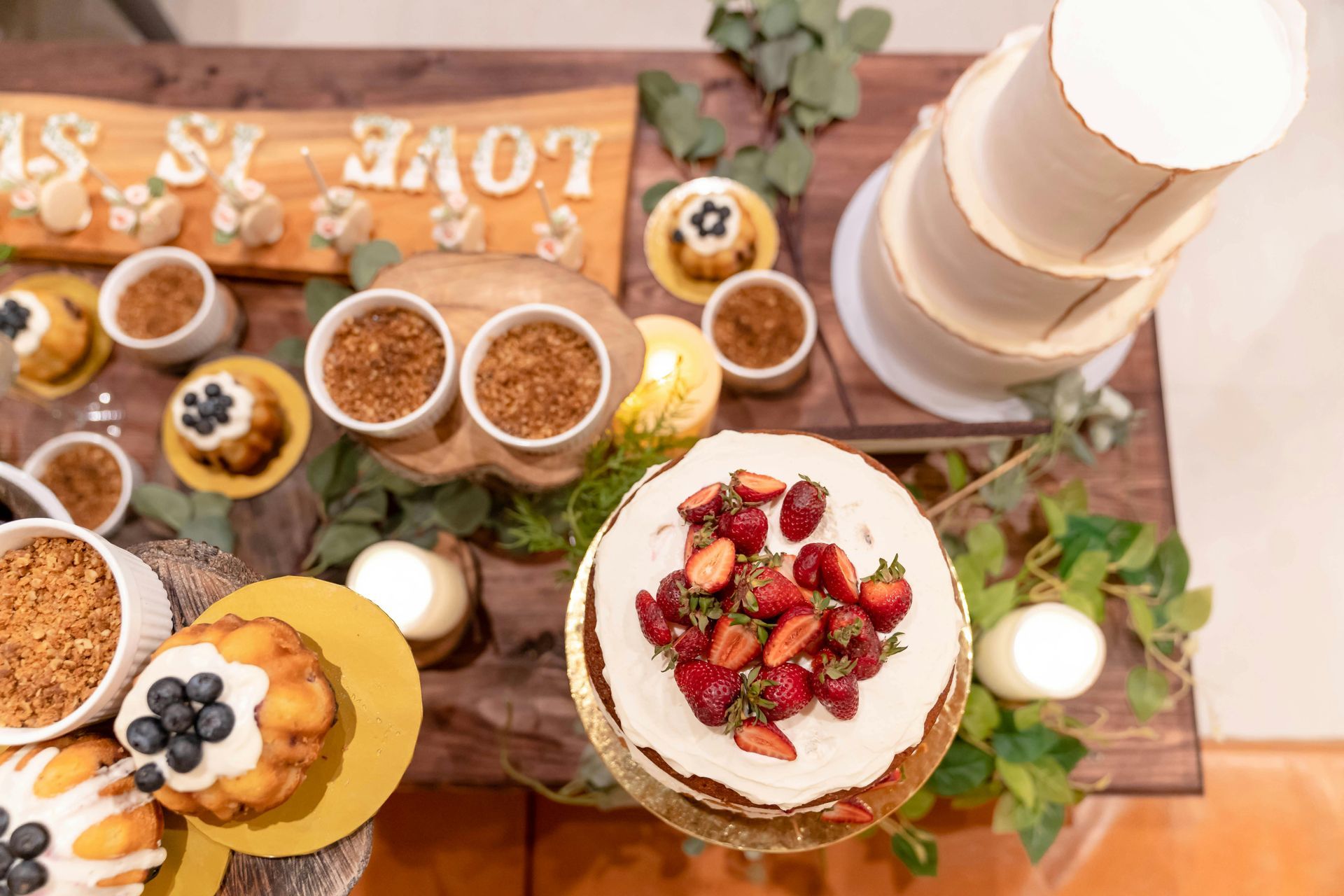 Dessert table with tiered cake, cupcakes, bowls of crumble, and a cake decorated with strawberries.