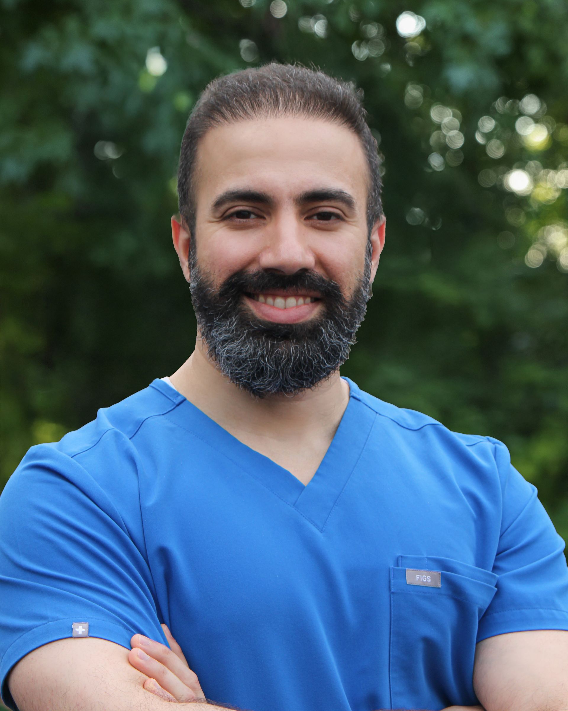 Man with dark hair and beard, wearing blue scrubs, smiling with arms crossed, outdoors.