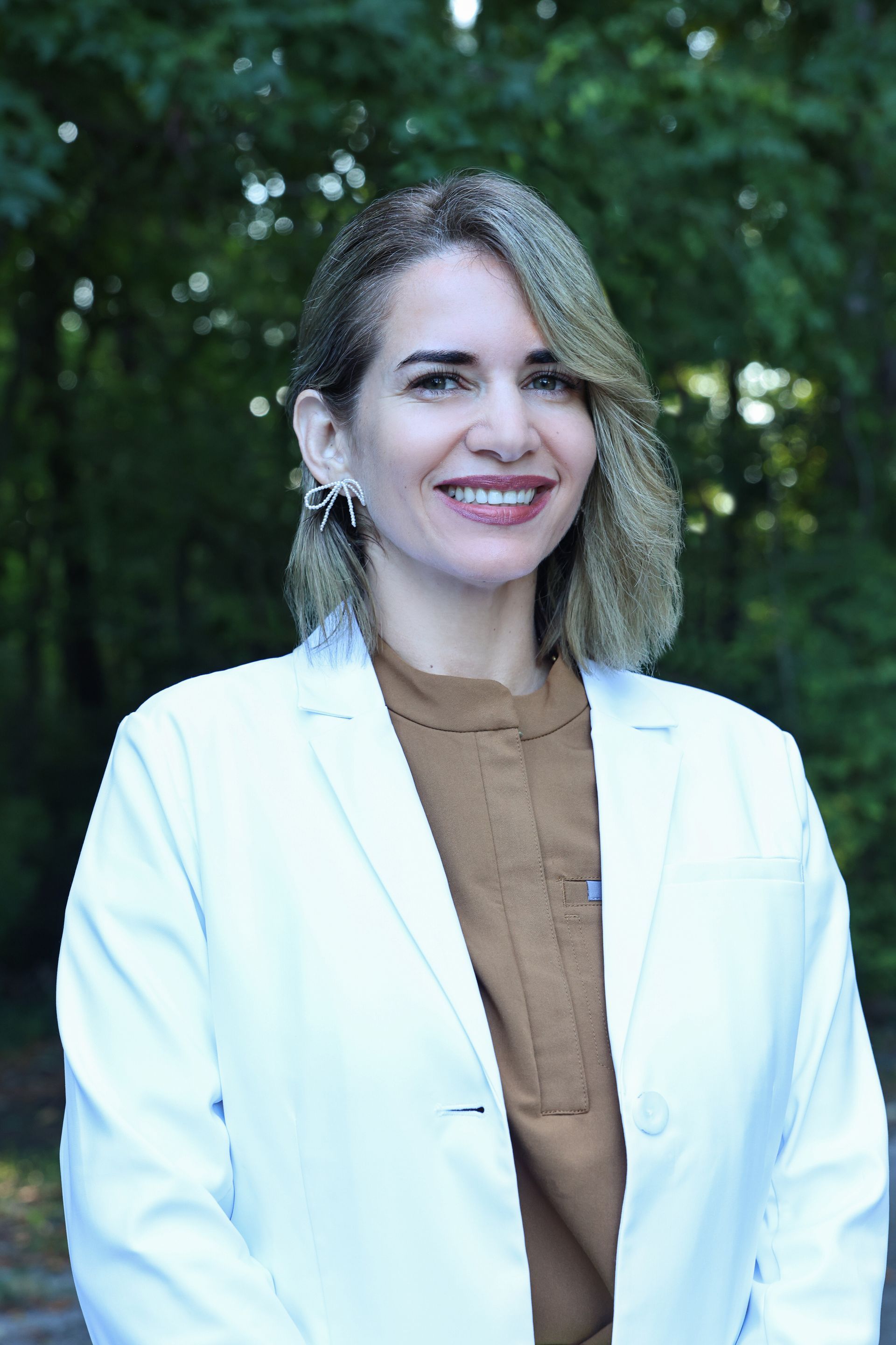 Woman in a white coat smiles outdoors; blurred green background.