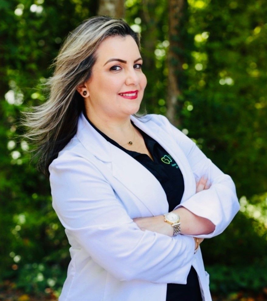 Woman in white coat, arms crossed, smiling, with a natural background.