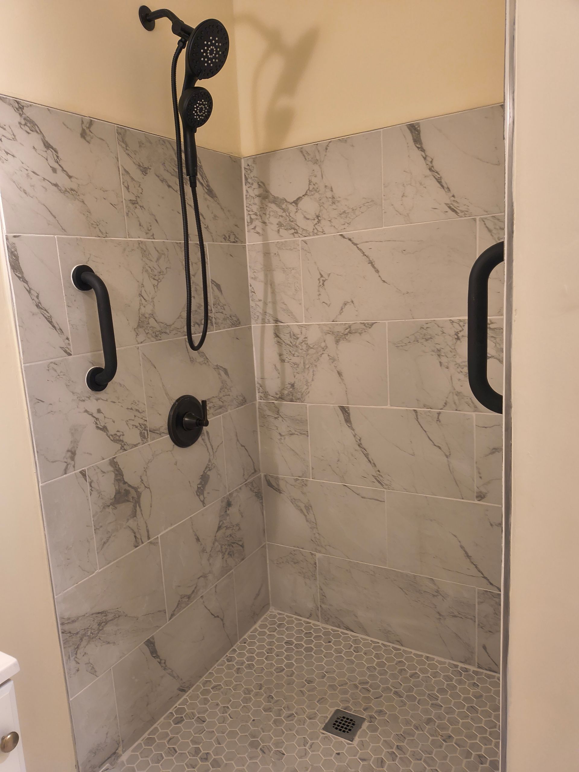 Marble-tiled walk-in shower with black grab bars and handheld showerhead.