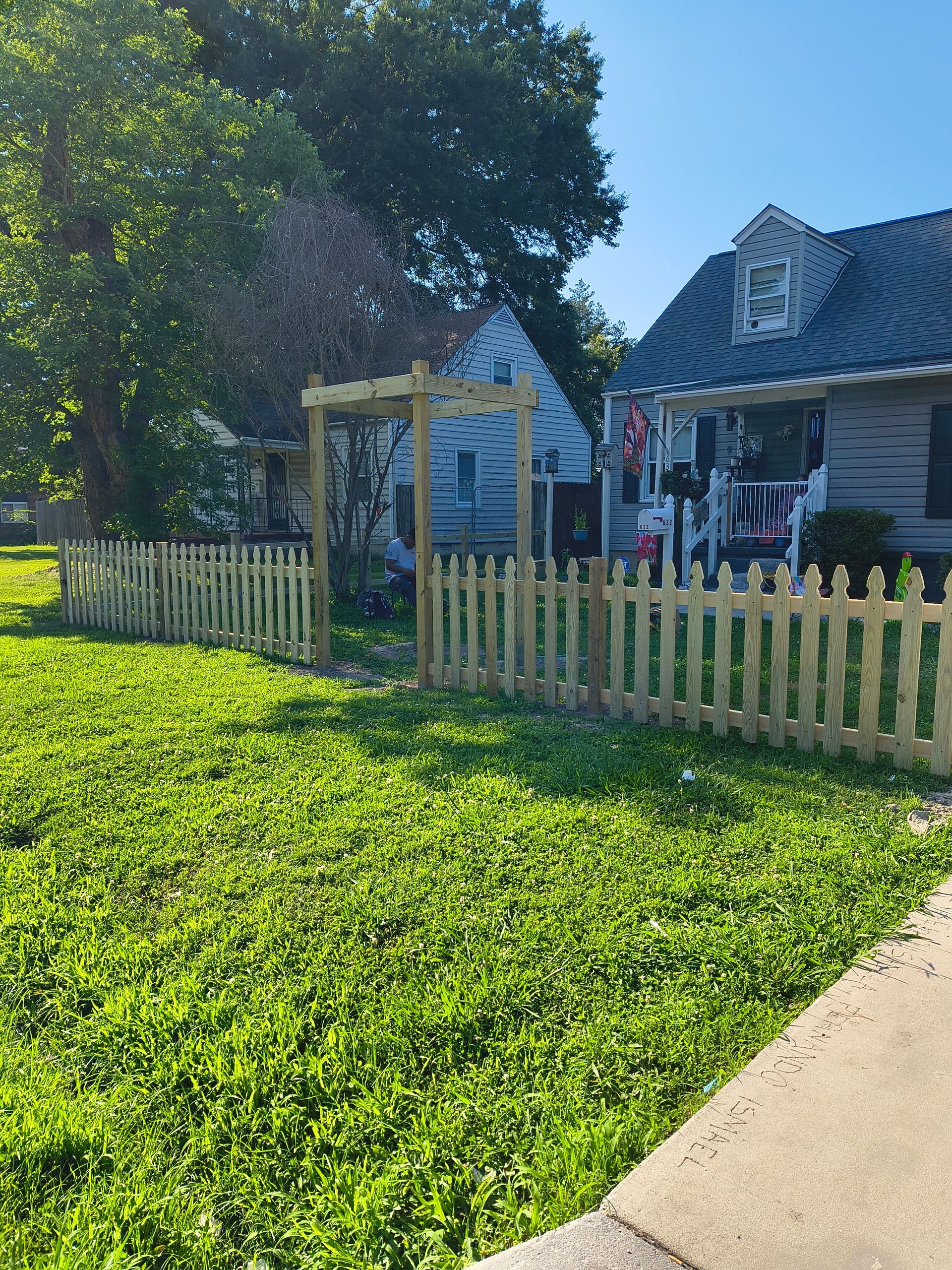Sunny front yard with green lawn, picket fence, arbor, and house porch under a blue sky