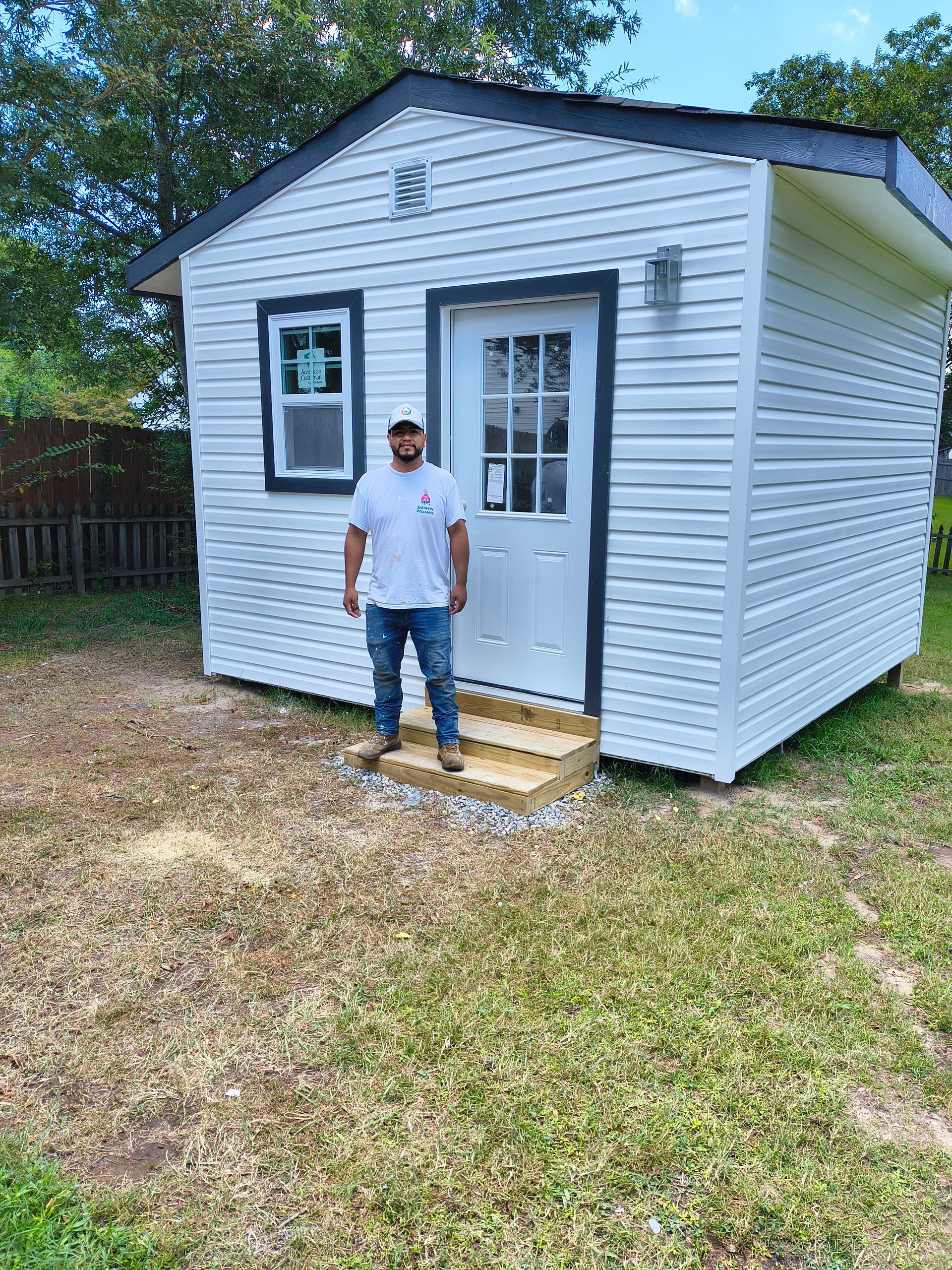 Person standing in front of a small white shed with a wooden step outside