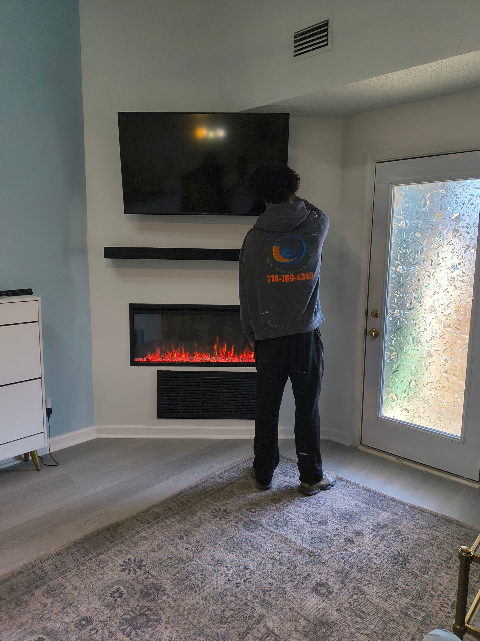 Person standing in a living room facing a wall-mounted TV and electric fireplace near a frosted glass door