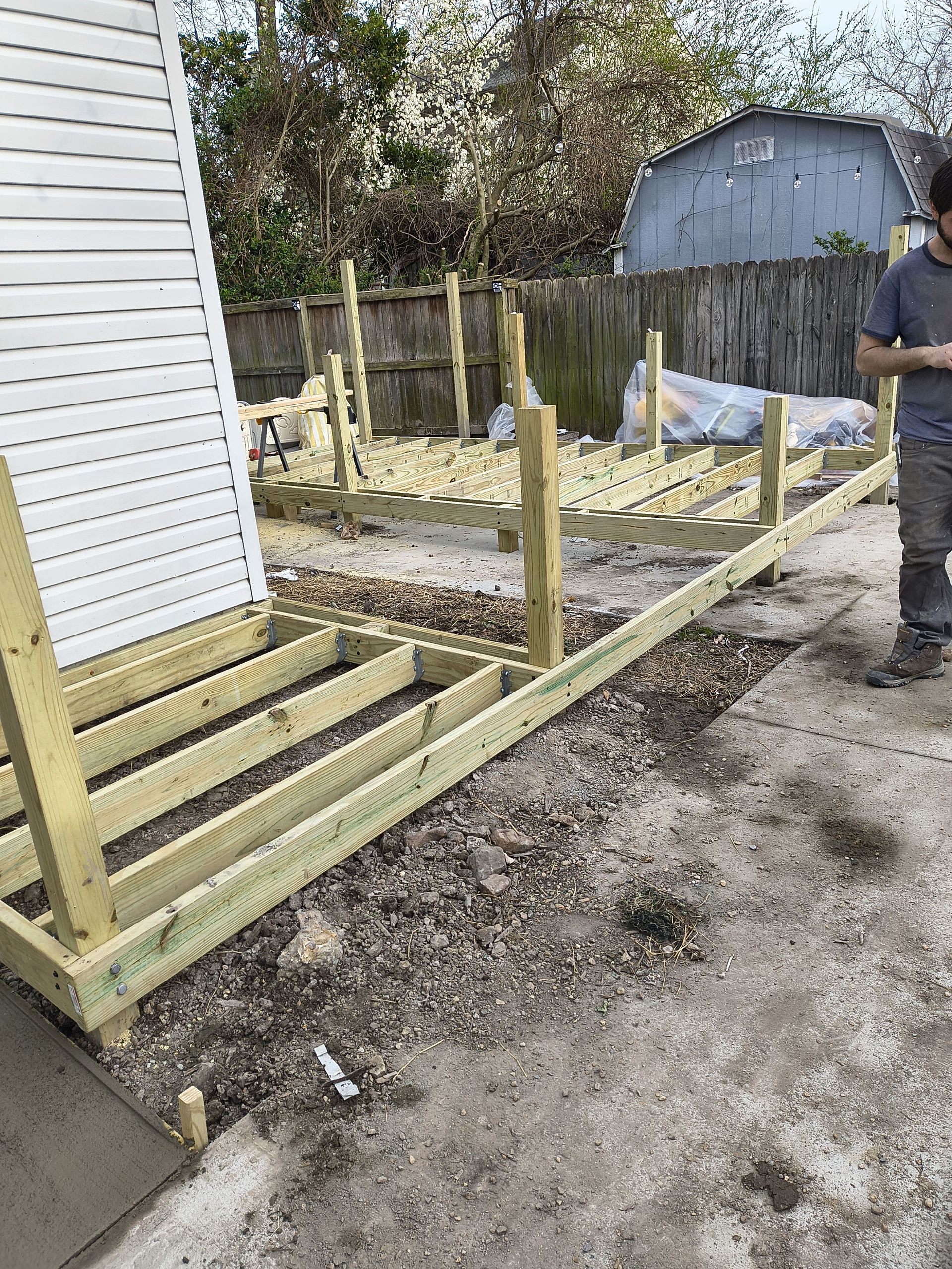Partially built wooden deck beside a house, with posts, railings, and a person standing nearby outdoors.
