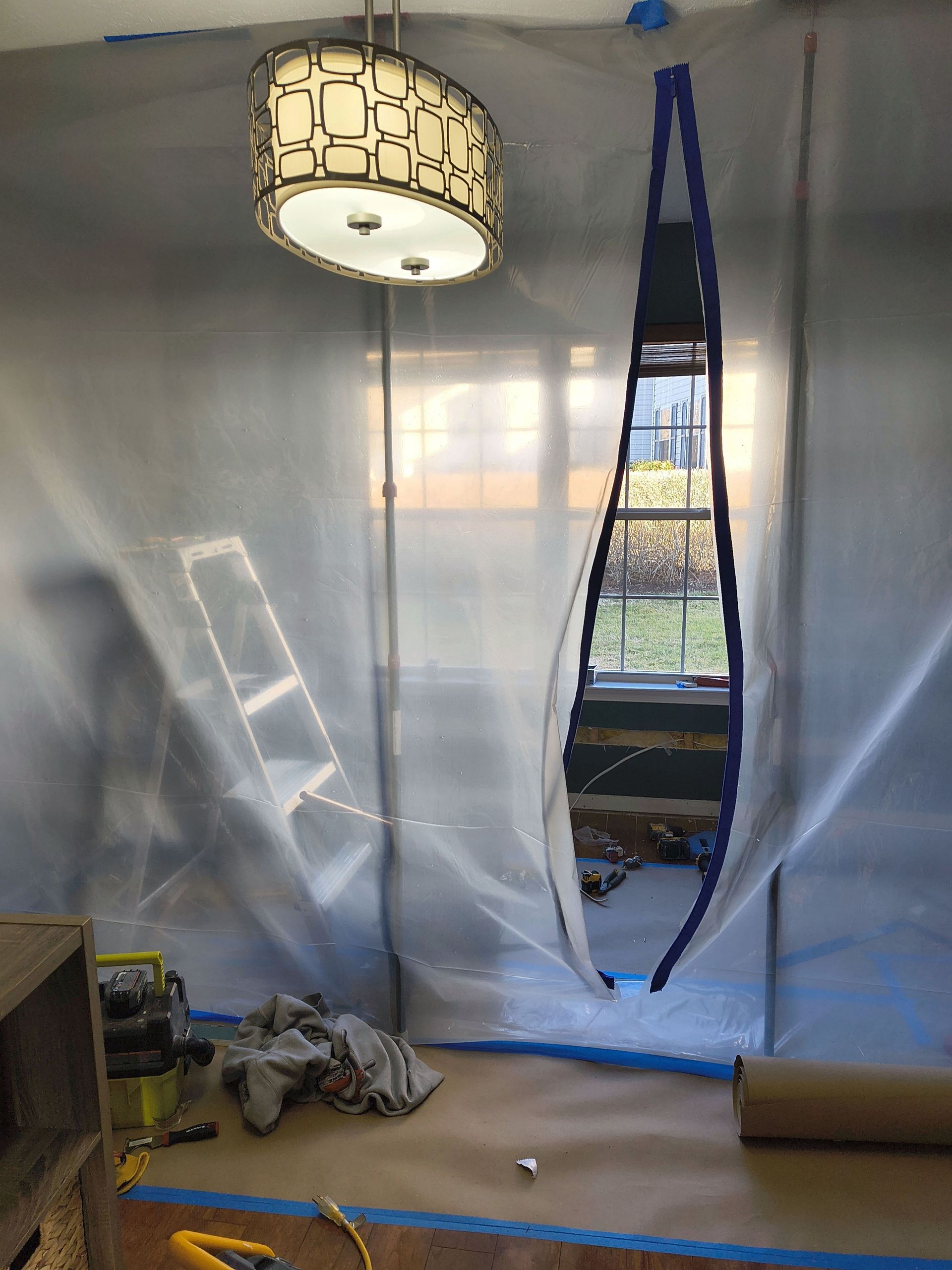 Plastic sheeting partitions a room with a window cutout, a hanging lamp, and scattered renovation tools.