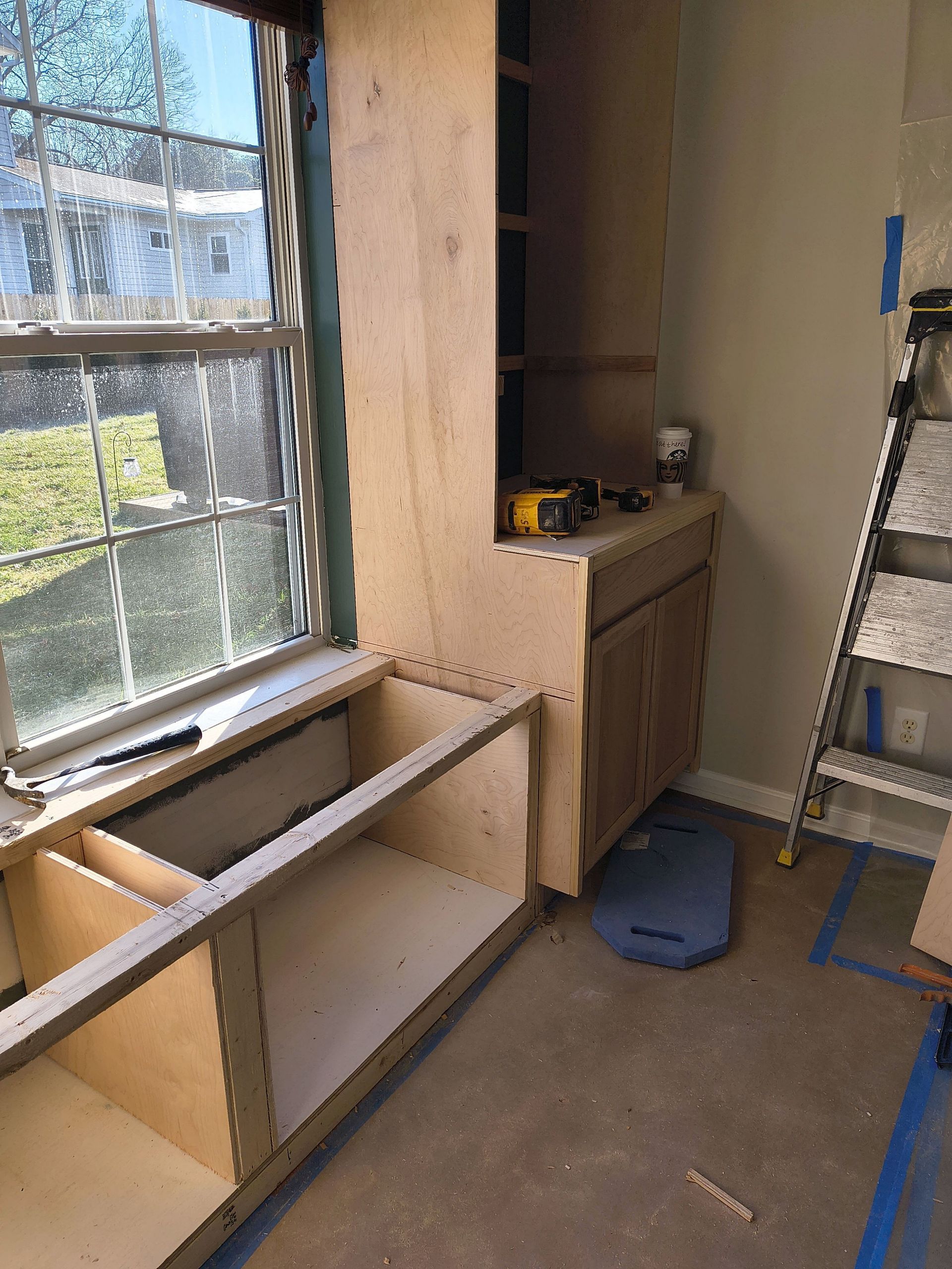 Partially built wooden cabinet in a room by a window, with a ladder and tools nearby.