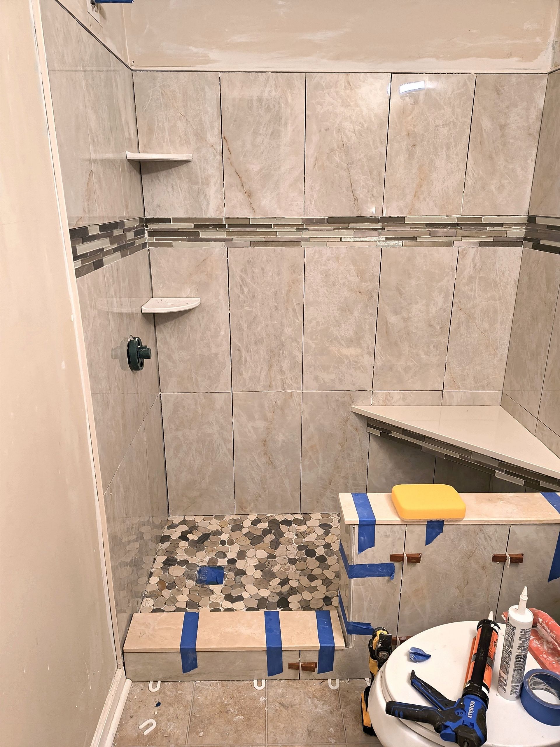 Partially tiled shower under renovation with exposed walls, blue tape, and construction tools on a shelf.