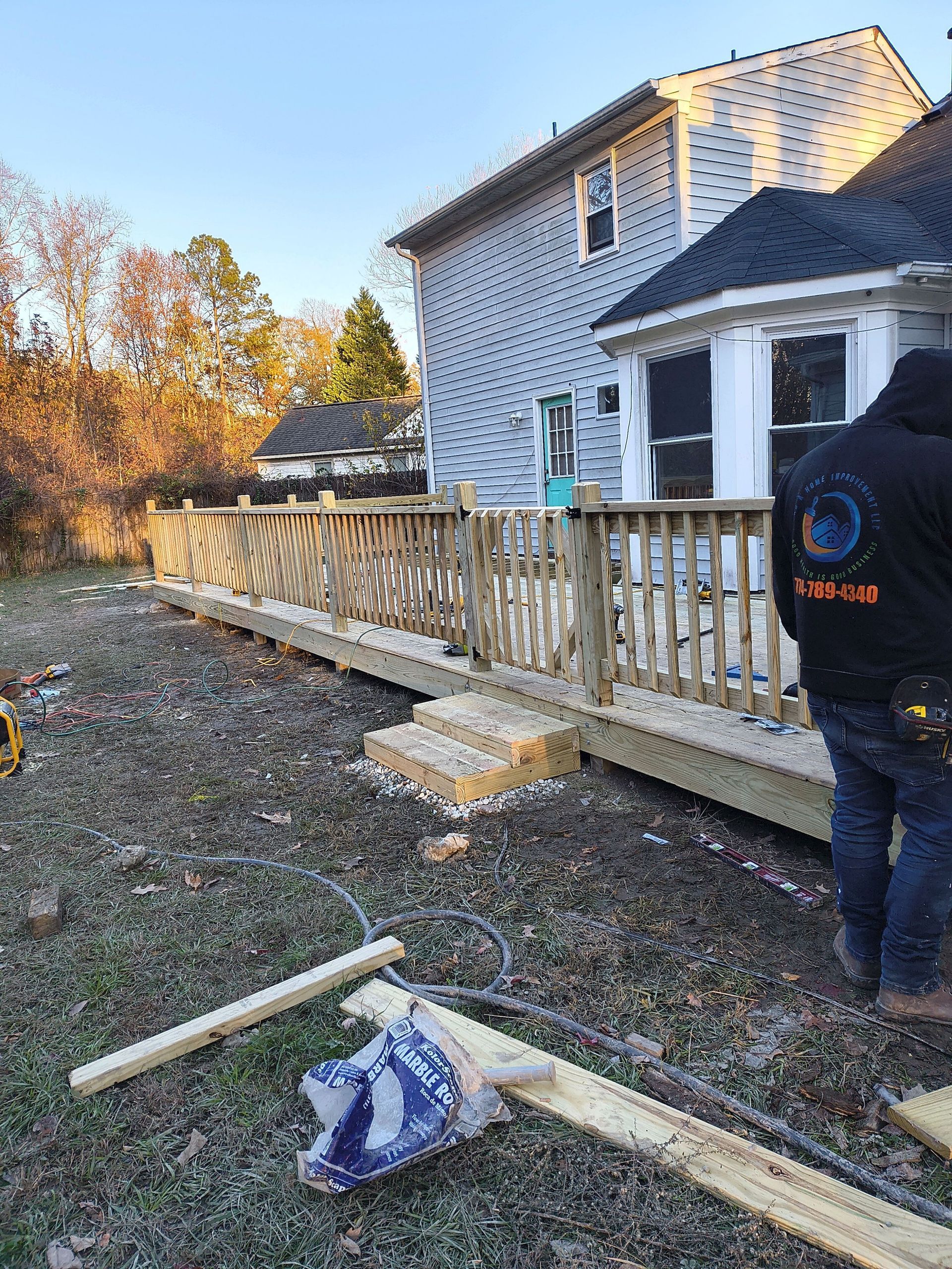 Person by a white house deck, with lumber and tools on the ground in a yard at dusk.