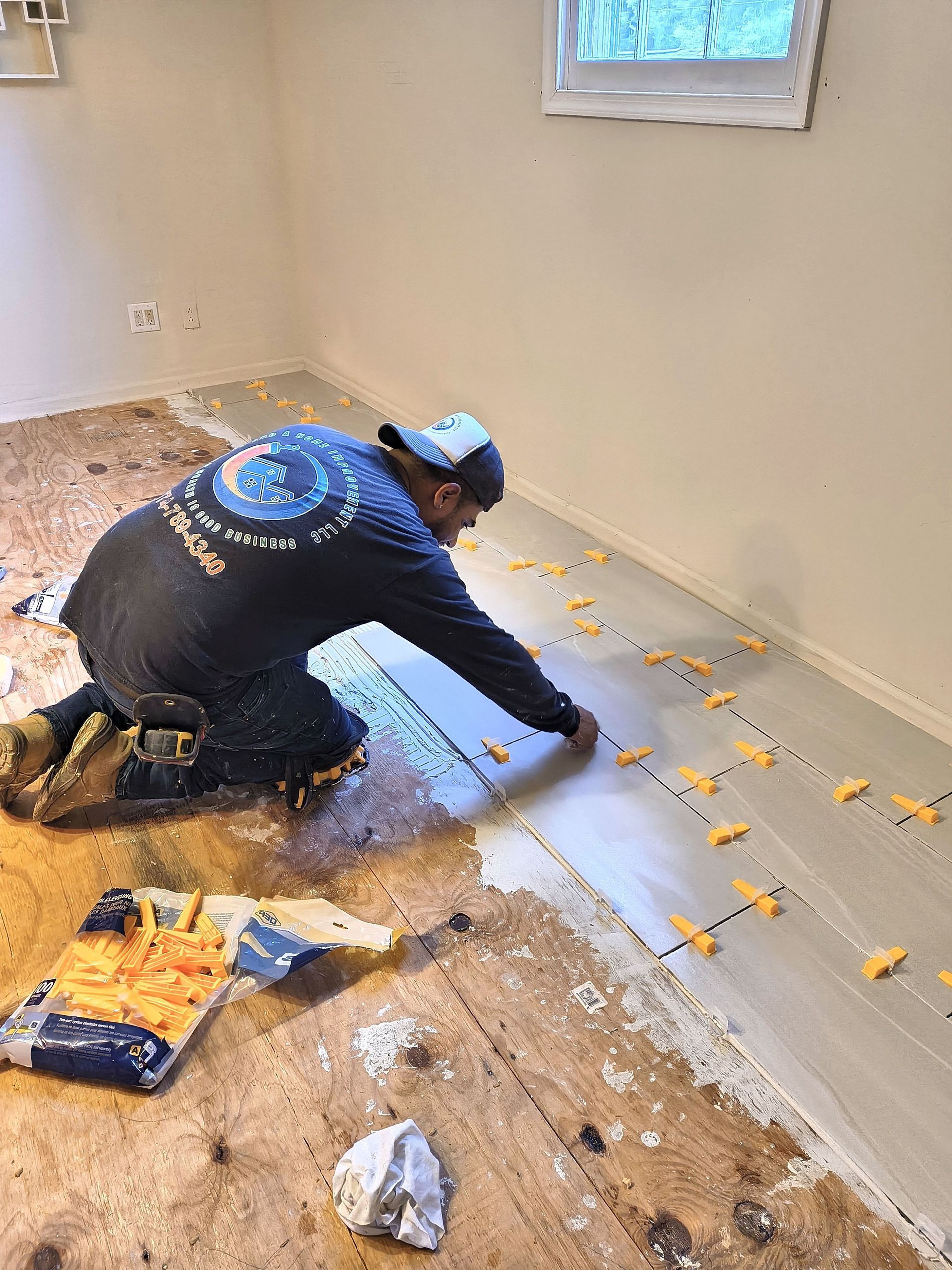 Worker laying floor tiles in a small room with white walls and a window.