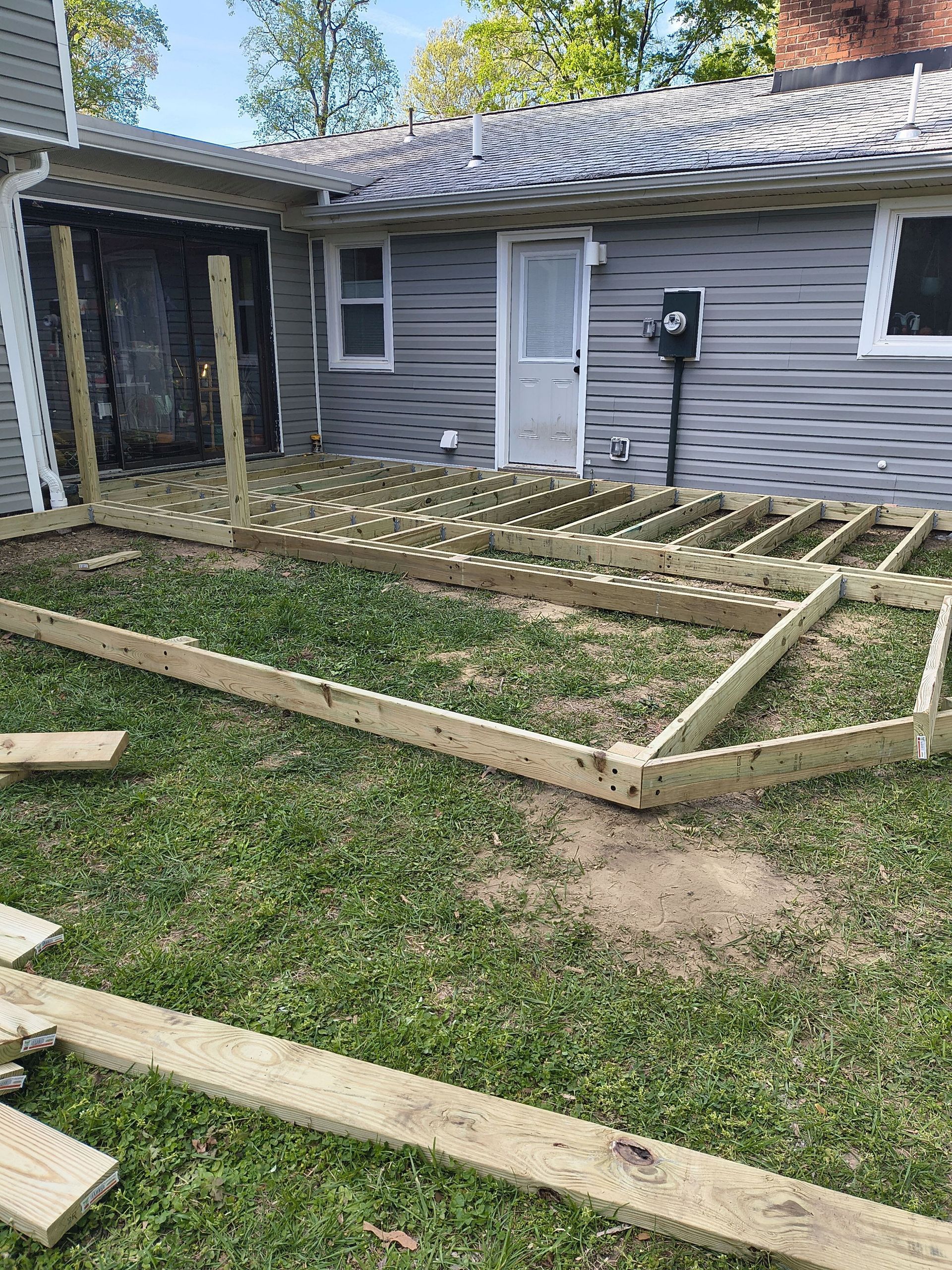 Partially built wooden deck frame beside a gray house with a lattice-covered porch and lawn.