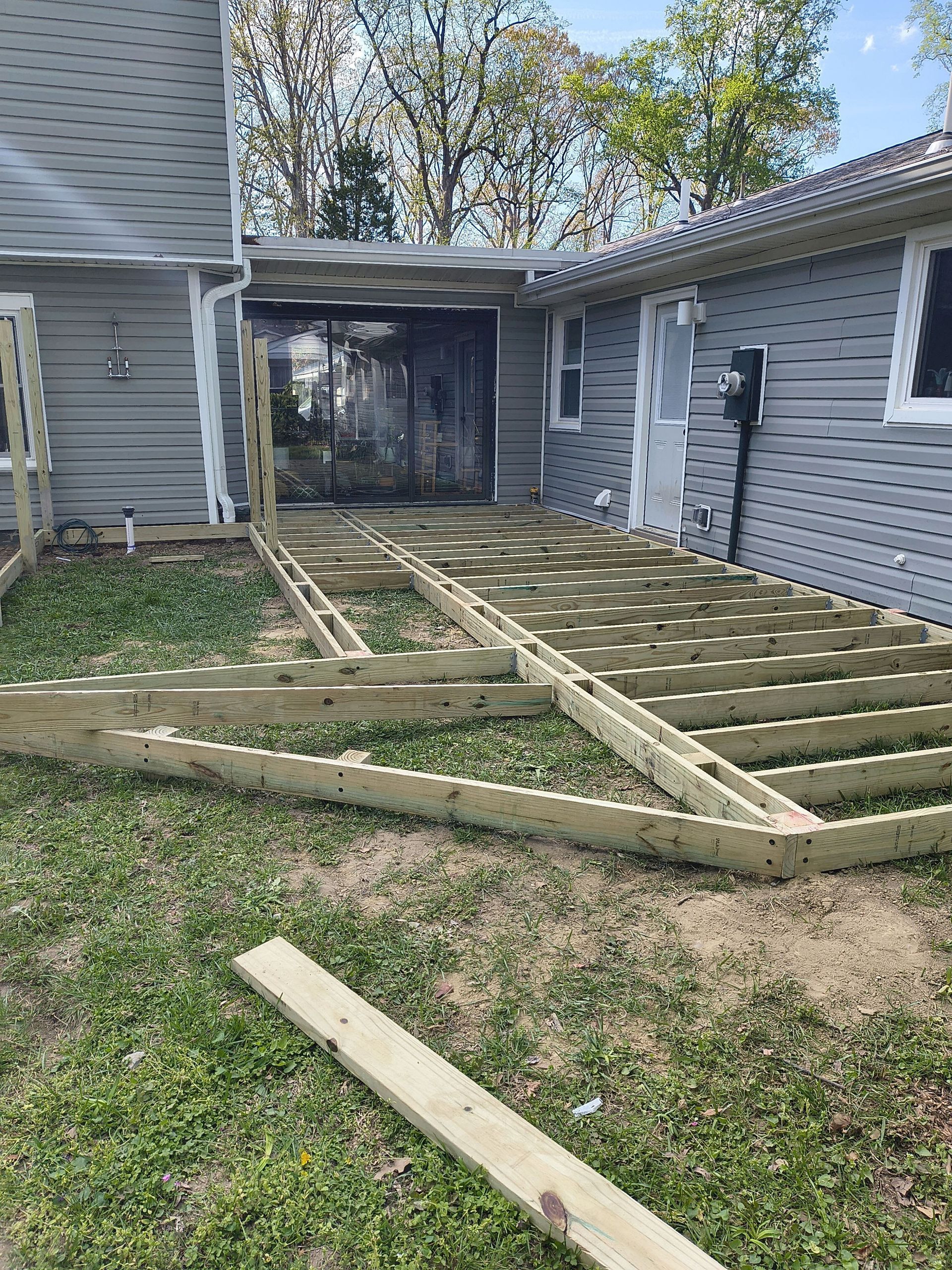 Partially built wooden deck framing in a backyard beside a gray house.