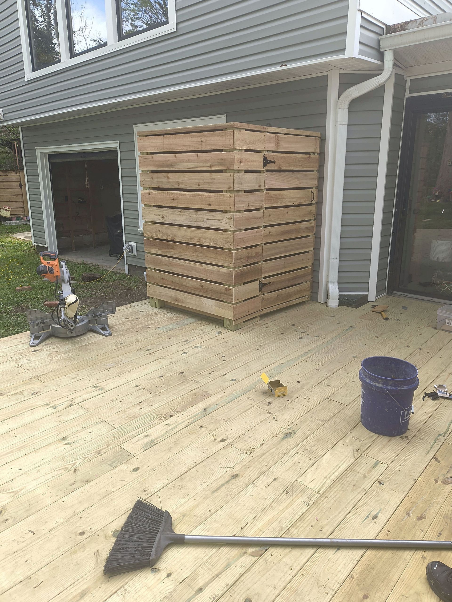 Wooden shed under construction beside a gray house, with stacked lumber and a broom on a deck.
