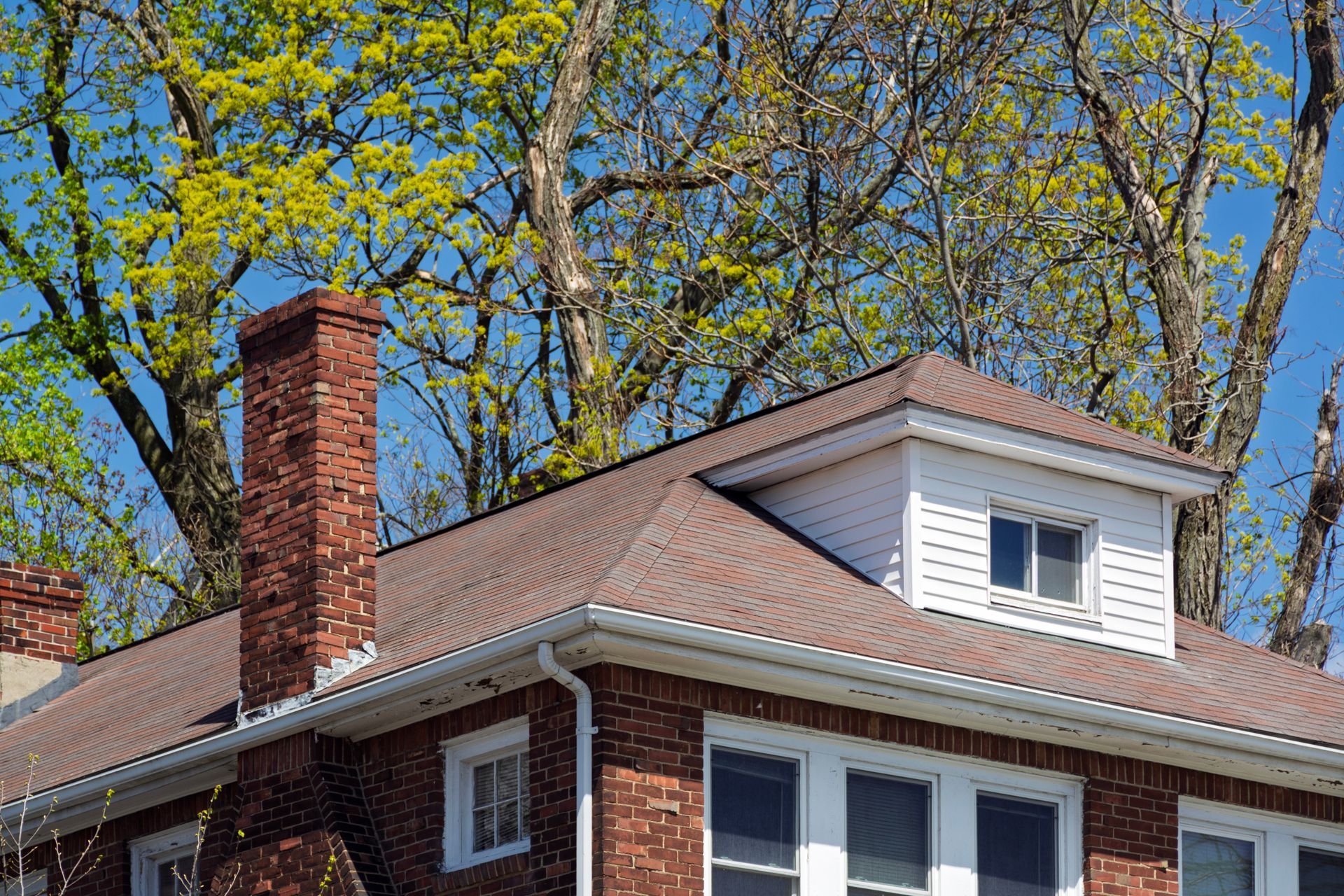 Brick house roof with red chimney and dormer window under leafy trees Brick house roof with red chimney and dormer window under leafy trees