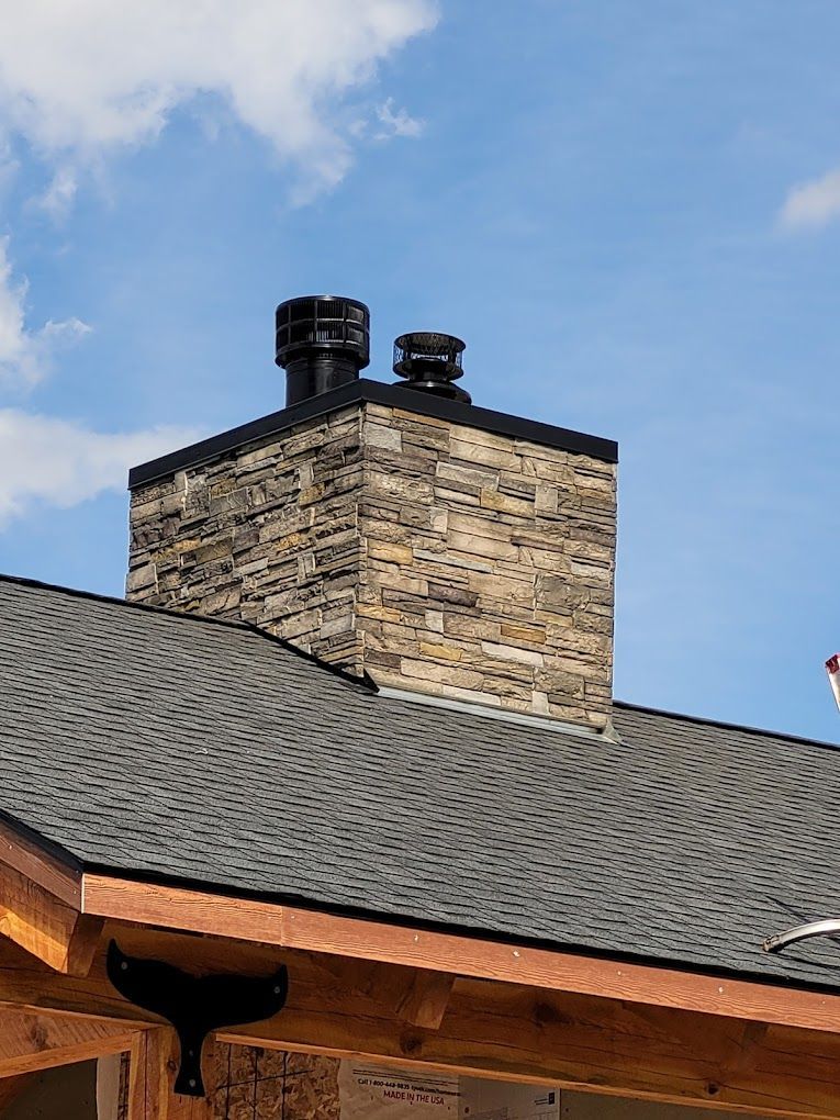 Stone chimney with black caps on a gray shingled roof, against a blue sky.