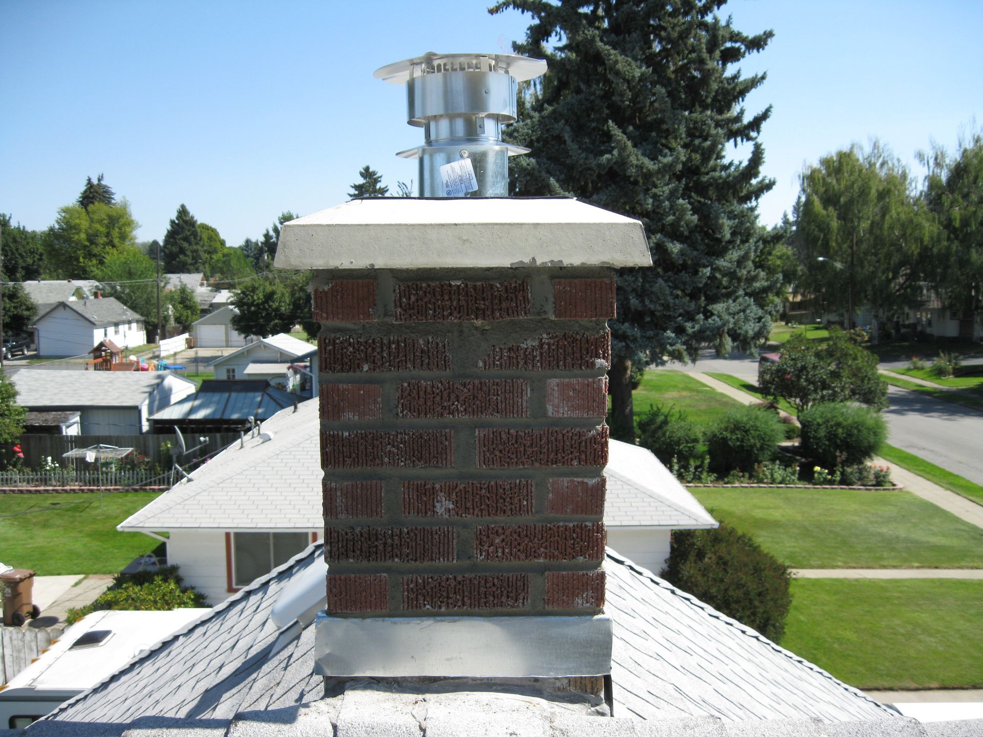 Brick chimney on rooftop with cap. Houses and trees in the background under blue sky.