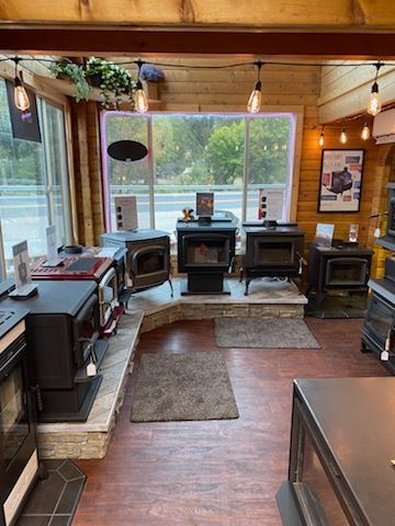 Wood stove showroom with multiple models on display, natural wood paneling, large windows, and overhead lights.