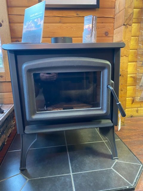 Black wood-burning stove with glass door, sitting on a tile floor in front of a wooden wall.