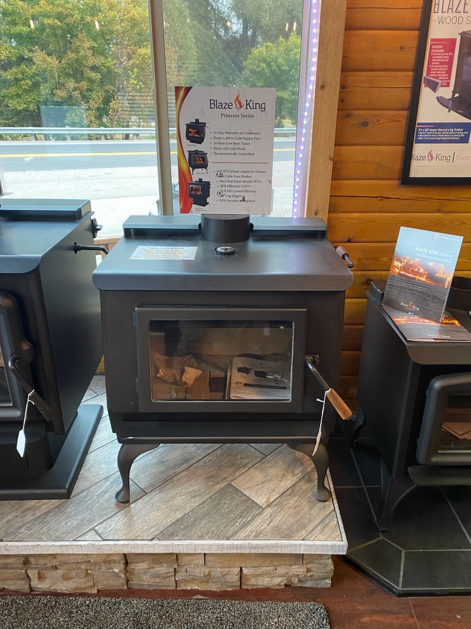 A wood-burning stove with glass door displayed inside a store, near a window.