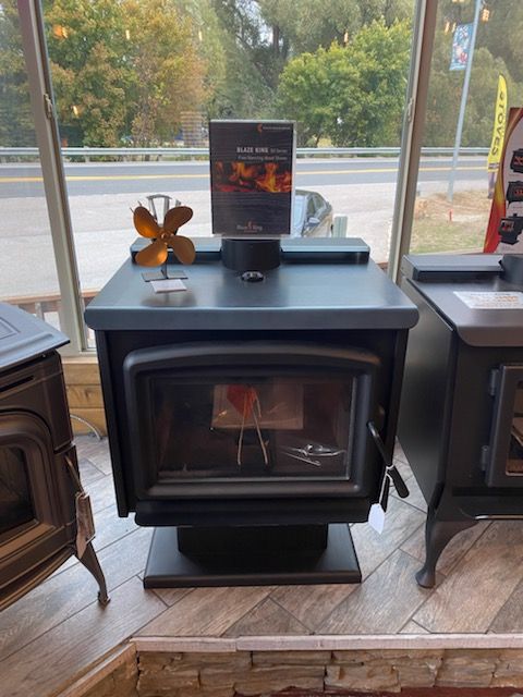 Black wood-burning stove with glass door and decorative fan on top, in a showroom window.