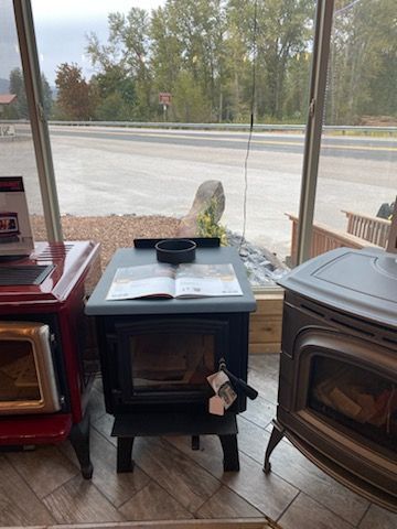 A black wood stove on display in a store, with a window view of a road and trees.