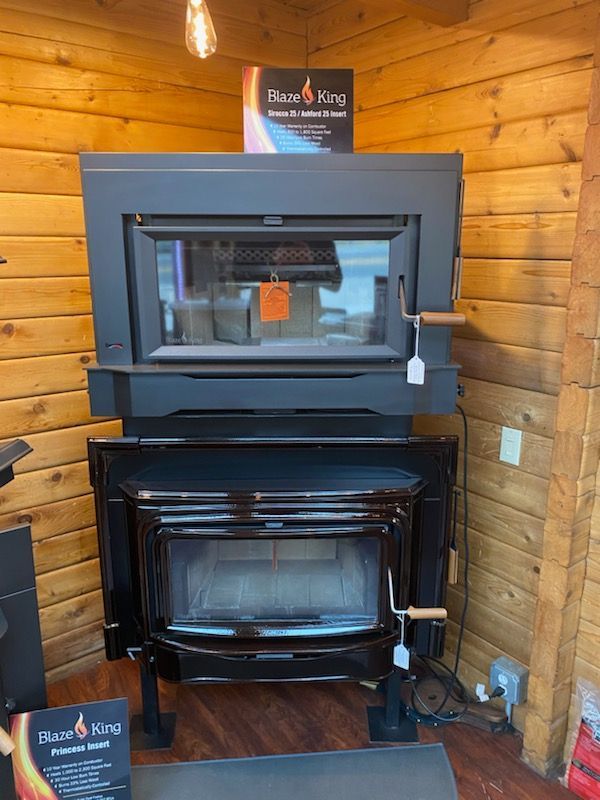 Two black Blaze King wood stoves in a wood-paneled room, one inset and one freestanding.
