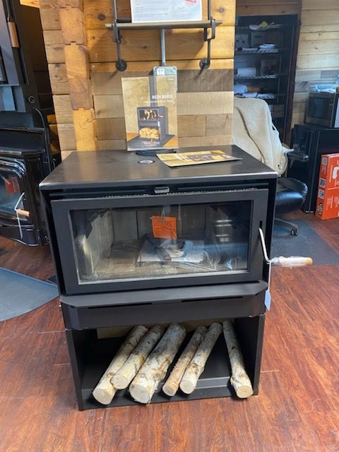 Wood-burning stove with glass door, firewood storage below. Displayed indoors with wooden paneling.