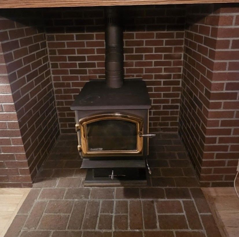 Wood-burning stove in brick fireplace. Brown stove and chimney against red brick backdrop. Wooden mantle above.