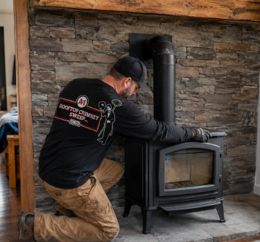 Person kneeling beside a black wood stove in a stone fireplace, adjusting it with gloved hands. Person kneeling beside a black wood stove in a stone fireplace, adjusting it with gloved hands.