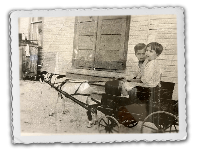 Two people sit in a small cart pulled by a goat in front of a wooden building, in a vintage black-and-white photograph.
