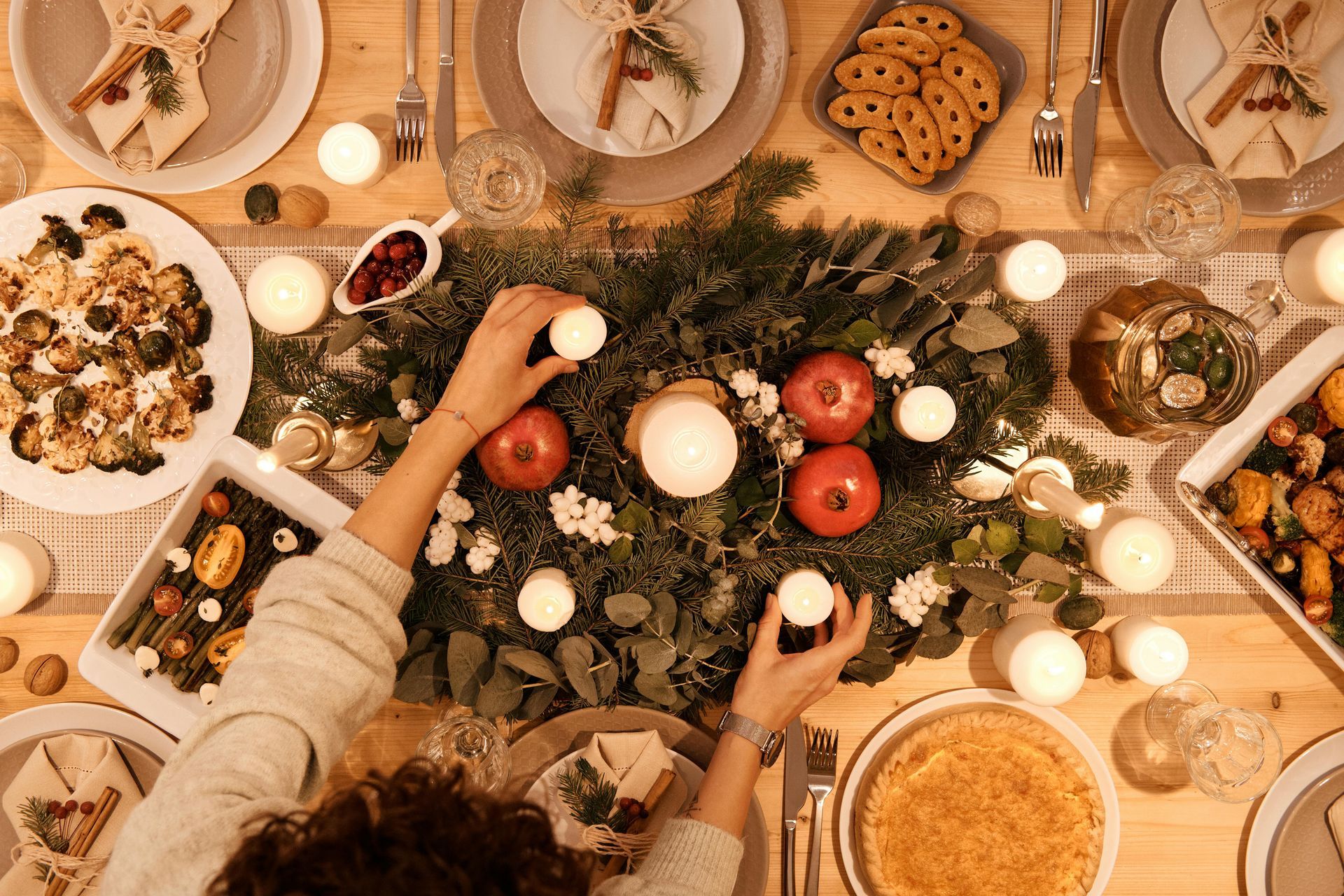 Overhead view of a decorated holiday table with a person lighting candles amidst food and greenery.
