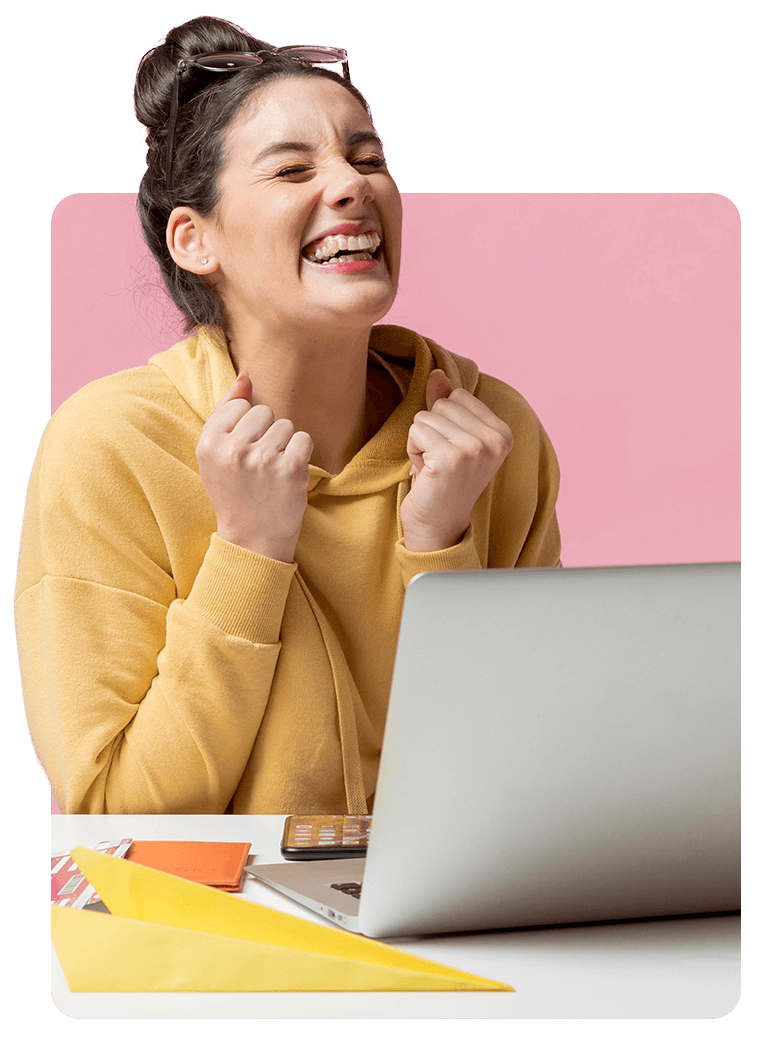Mujer con sudadera amarilla, con los brazos levantados, sonriendo frente a una computadora portátil sobre un escritorio blanco, fondo rosa.