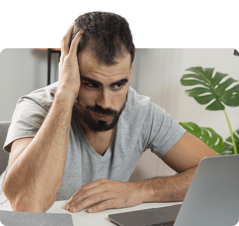 Hombre mirando estresado a una computadora portátil, con la mano en la cabeza, en el interior con una planta.