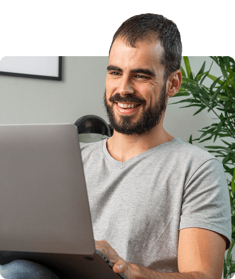 Hombre con barba sonriendo, trabajando en una computadora portátil en el interior con plantas.