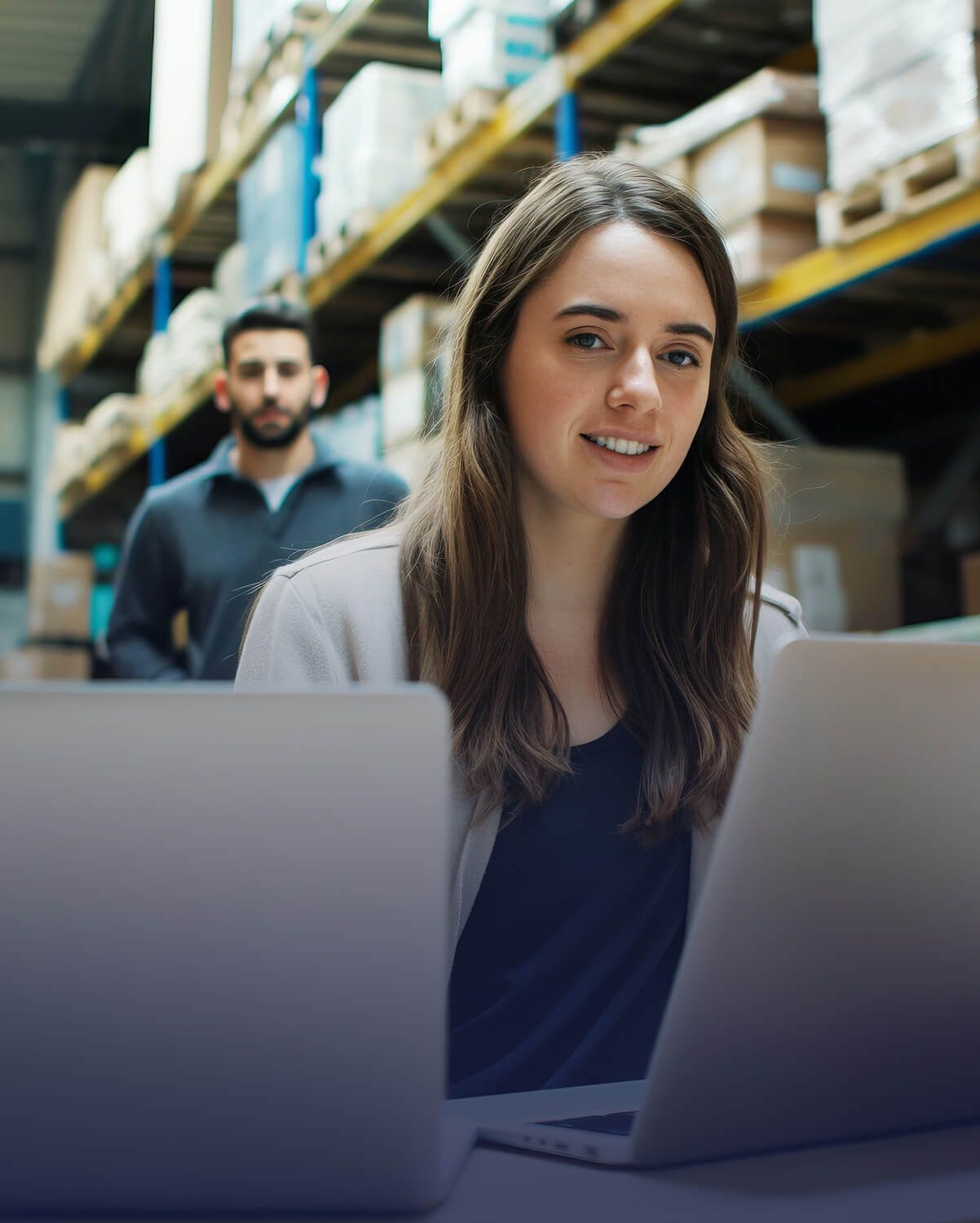 Mujer con portátil sonriendo en un almacén, otra persona caminando en el fondo.