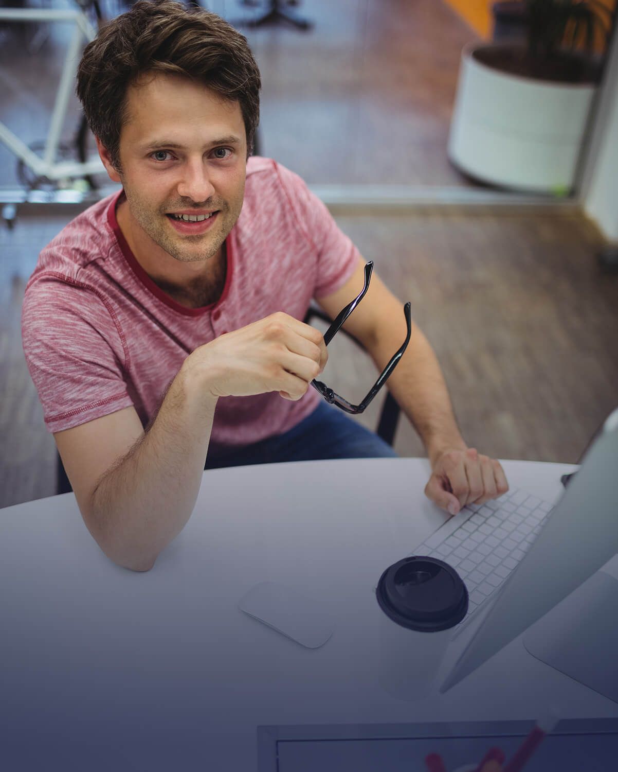 Un hombre sentado en un escritorio con gafas, sonriendo a la cámara. Está en un espacio de trabajo informal.