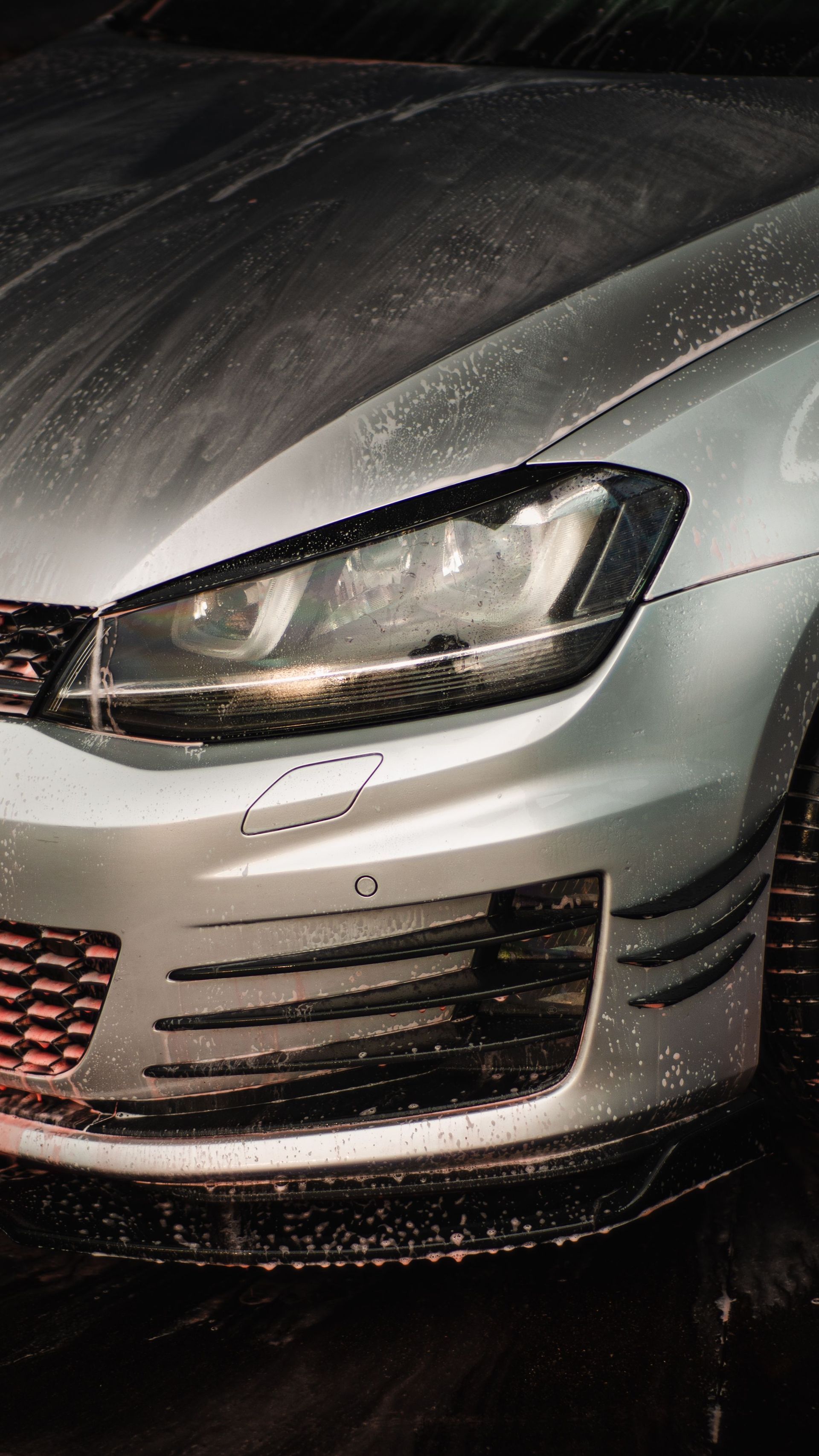 Close-up of a silver car’s front end with a headlight and grille at night, wet with rain.