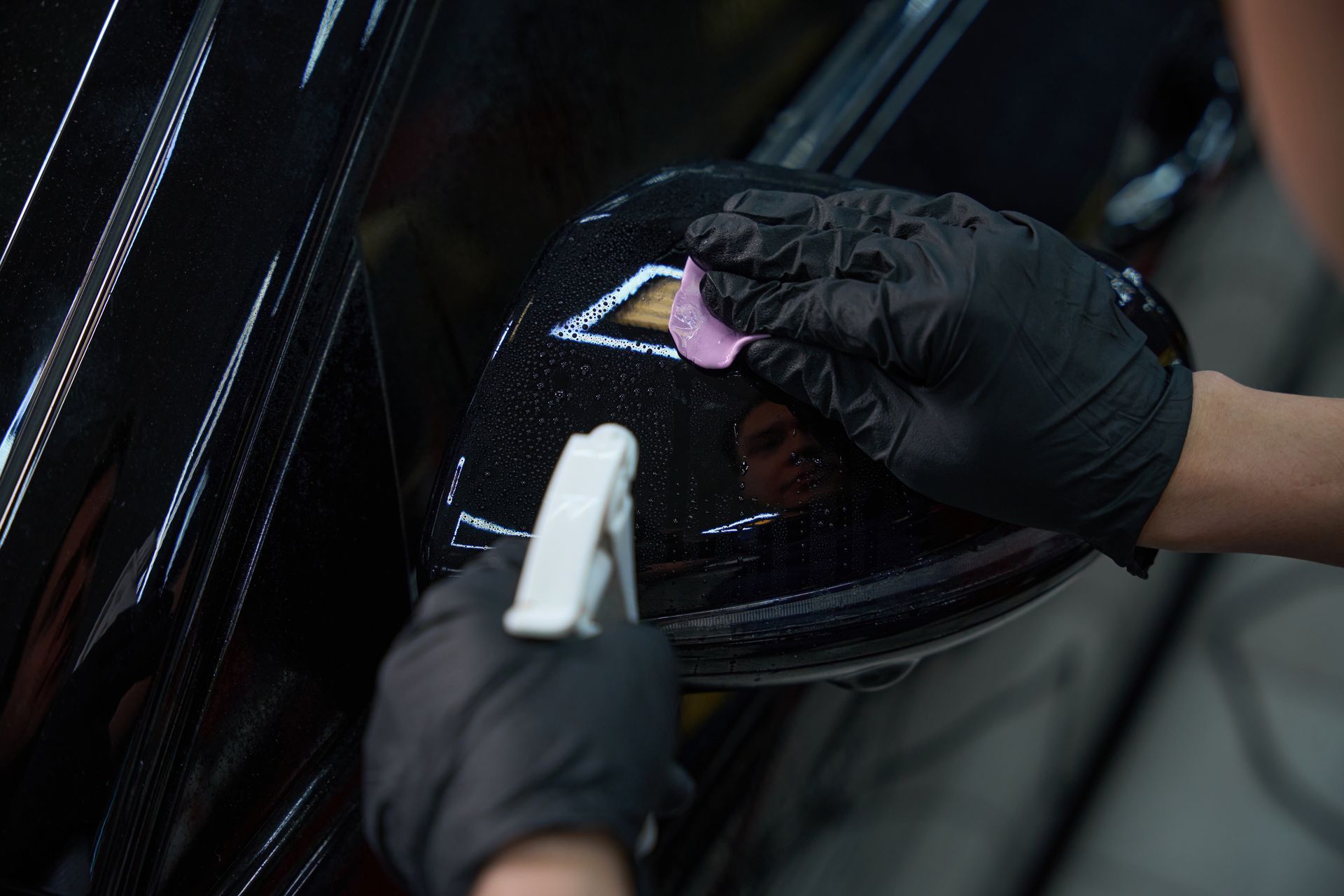 Gloved hands cleaning a car’s glossy black surface with a spray bottle and cloth