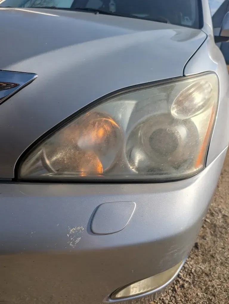 Close-up of a silver car’s front headlight and bumper with a small scuff mark.