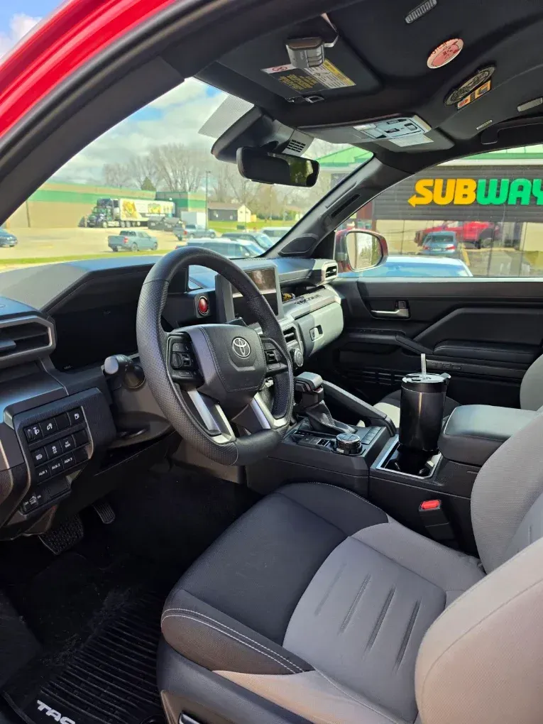 Driver’s seat view of a black-and-gray car interior with a steering wheel and center console, parked near a Subway restaurant.