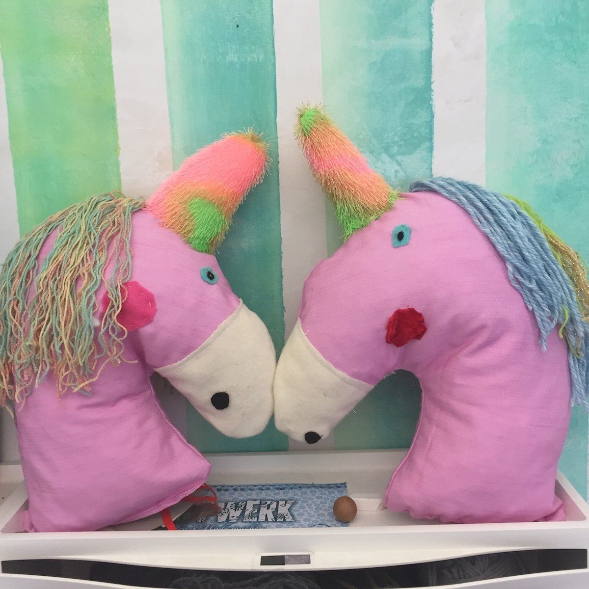 Zwei rosa Einhorn-Kopfkissen mit bunten Mähnen und Hörnern auf einem Regal.