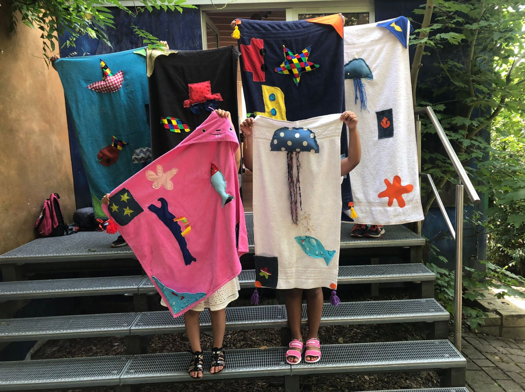 Zwei Kinder präsentieren Stoffkunstwerke vor einem Gebäude, im Hintergrund hängen weitere Stoffkunstwerke; Außenaufnahme.