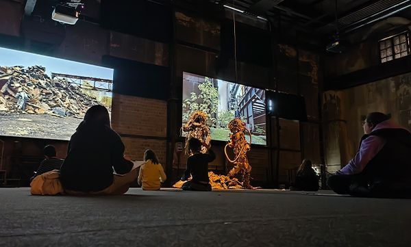 Menschen sitzen auf dem Boden und verfolgen eine Aufführung auf Großbildschirmen in einer Industriehalle.