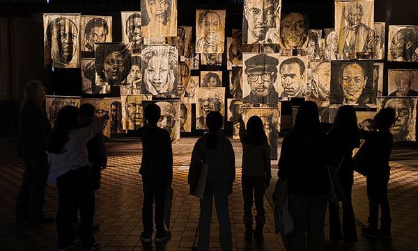 Menschen betrachten eine Galerie mit Kohleporträts an einer Wand, deren Silhouetten sich von den Kunstwerken abheben.