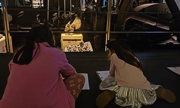 Zwei Personen sitzen und skizzieren auf Papier. Dunkle Museumsatmosphäre.