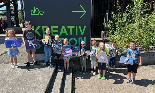 Eine Gruppe Kinder hält Kunstwerke vor einem Schild mit der Aufschrift „Kreativ Station“ hoch.