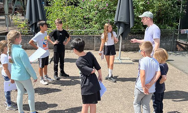 Kinder und ein Erwachsener sitzen im Freien im Kreis, einige halten Papiere in den Händen. Der Erwachsene gestikuliert, möglicherweise leitet er eine Aktivität an.